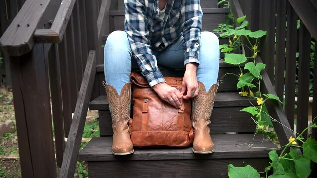 woman in blue jeans and cowboy boots unzips a leather backpack. Close-up.