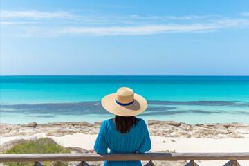 Fototapeta na wymiar Serene Woman in Stylish Hat Admiring Azure Sea and Sandy Beach on Exotic Coastal Getaway