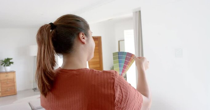 Thoughtful plus size biracial woman looking at paint colour charts in sunny living room, slow motion