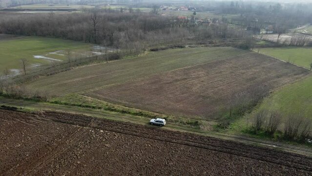Drone shot of car driving on dirt road surrounded by agricultural fields towards village