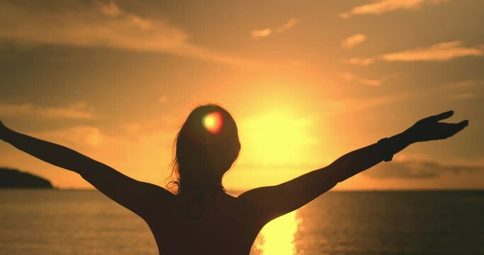 Woman portrait against sunset seascape bright orange sky. Girl silhouette raises hands ang enjoy beautiful nature landscape. Outdoor lifestyle travel on summer holiday vacation. Slow motion back view