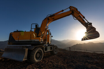 Excavator on the hill at sunset. Excavators are heavy construction equipment consisting of a boom, dipper (or stick), bucket and cab on a rotating platform. - obrazy, fototapety, plakaty