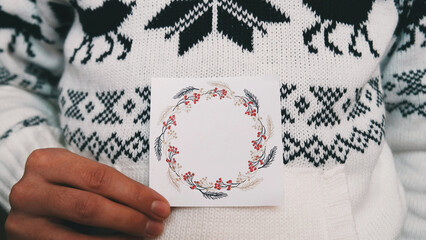 Woman in white embroidered cotton sweater holding up an empty Christmas card. - obrazy, fototapety, plakaty