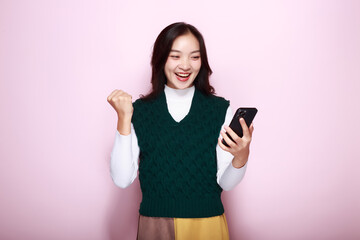 An Asian woman poses holding a phone and looks surprised while standing in front of a light pink...