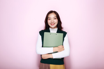 Fototapeta premium Asian woman poses holding a file folder while standing in front of a light pink background.