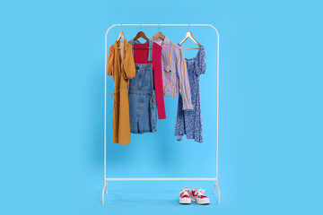 Rack with stylish women`s clothes on wooden hangers and sneakers against light blue background