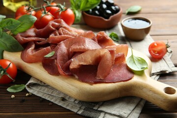 Board with delicious bresaola served with tomato and basil leaves on wooden table, closeup