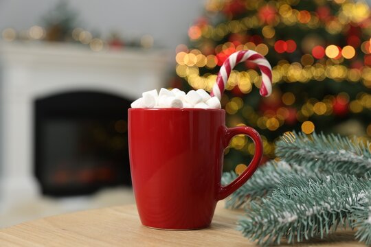 Christmas cocoa with marshmallows and candy cane in red cup on wooden table indoors, closeup