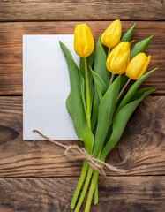 Top view of a bouquet of yellow tulips and a piece of paper for text on a wooden background with copy space
