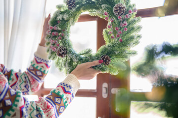 Young asian woman in sweater with happiness hanging and decorating christmas wreath on the door while preparing decoration ornaments to celebrate for xmas festive holiday and winter seasons at house