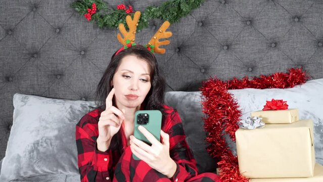 Young beautiful woman in red plaid pyjama borred while sliding on smartphone in Christmas decorated room with presents.