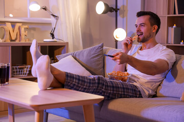 Young man with potato chips watching TV at home in evening