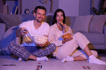 Young couple with potato chips and popcorn watching TV at home in evening
