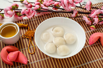 Bamboo mat with bowl of tangyuan, tea, fortune cookies and sakura on white marble background,...