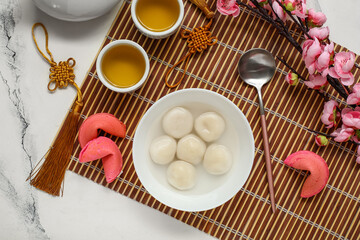 Bamboo mat with bowl of tangyuan, tea, fortune cookies and sakura on white marble background. Dongzhi Festival