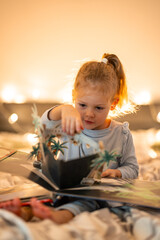 Fototapeta premium Little girl reading pop up book in home bed in Christmas environment with lights background