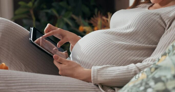 Pregnant woman in a close-up shot, holding a tablet in her hands, scrolling through photos with a focused look touching belly .