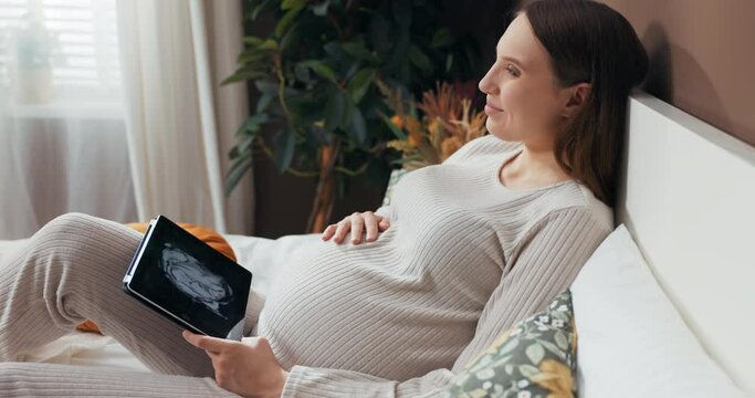 Beautiful pregnant woman in her late 20s to early 30s, dressed in a fashionable and cozy outfit, sitting on a bed, and flipping through photos on a tablet.