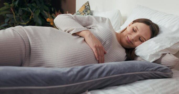 Smiley pregnant woman in a comfortable outfit, lying on a pregnancy pillow, and lovingly caressing her belly while beaming with happiness.