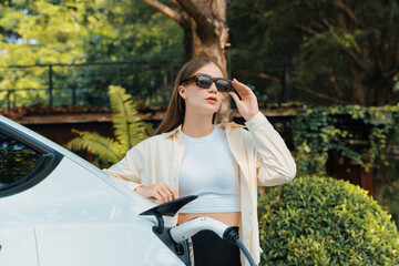 Young woman recharge her EV electric vehicle at green city park parking lot. Urban sustainability...