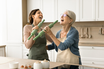 Joyful older mom and excited adult daughter having fun in kitchen, acting singers while baking pastry, singing song at roller mics, shouting, laughing, having fun, enjoying family cooking, friendship - Powered by Adobe