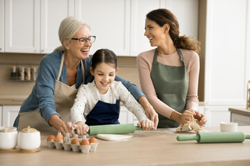 Obraz na płótnie Canvas Cheerful grandma teaching little granddaughter kid and adult daughter woman to bake in home kitchen, rolling dough for preparing homemade bakery food, talking, laughing, having fun