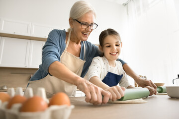 Positive baker grandma helping cute granddaughter kid to roll dough in home kitchen. Happy...