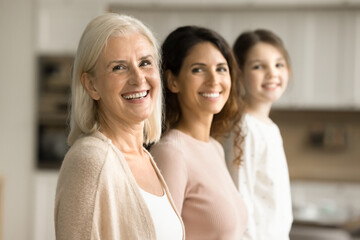 Happy attractive blonde senior grandma standing in row with adult daughter woman and little granddaughter kid, looking at camera with toothy smile, posing for three generations family portrait