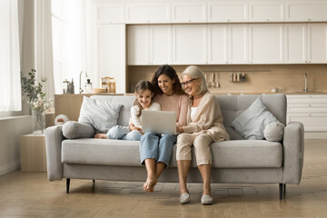 Cheerful elderly grandma, young mother and sweet little kid girl using laptop computer, enjoying wireless technology, domestic Internet communication, resting on couch in comfortable home interior - Powered by Adobe