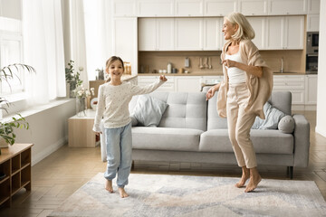 Cheerful granddaughter child and happy active blonde grandma dancing to music in cozy interior, smiling, laughing, having fun at home party, enjoying active leisure, playtime