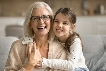 Happy pretty tween granddaughter girl hugging loving grandma from behind, looking at camera,...