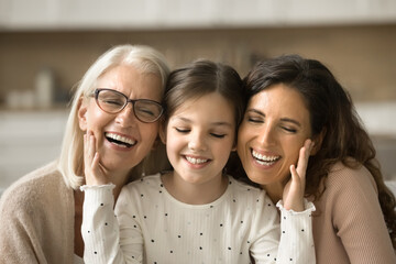 Happy little kid girl touching soft facial skin of pretty grandma and mother. Cheerful granny, mom...
