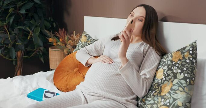 Beautiful pregnant woman lying on bed with photos in lap talking on phone with smile on face photos newborn baby woman may be discussing plans updates related to pregnancy.