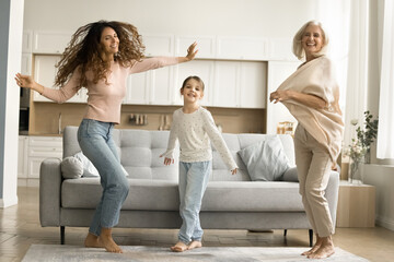 Happy kid girl, excited mom and cheerful granny dancing together in living room, laughing, singing...