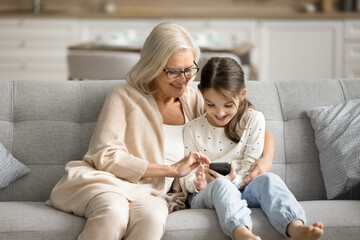 Cheerful grandmother and happy tween kid girl using application, Internet service on mobile phone,...