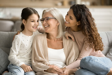 Cheerful grandma, mom and grandkid girl resting on home couch, talking, laughing, having fun,...