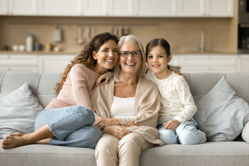 Happy grandma, mom and granddaughter kid sitting close on home sofa. Young mother and girl hugging...