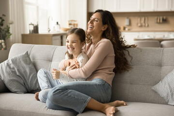 Happy carefree mom tickling cheerful daughter kid on sofa, cuddling cute child, laughing, enjoying...