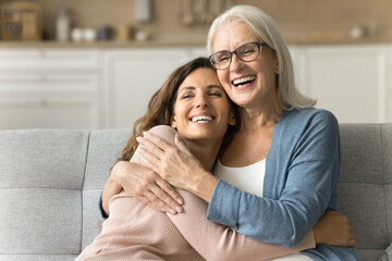 Cheerful attractive senior mom and happy adult daughter hugging at home with love, care, resting on couch together, smiling, laughing, talking, enjoying family relationship, bonding, motherhood - obrazy, fototapety, plakaty