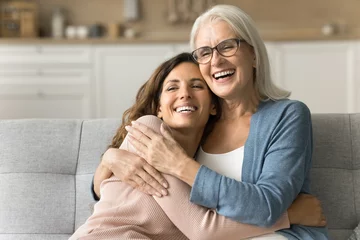 Fotobehang Cheerful attractive senior mom and happy adult daughter hugging at home with love, care, resting on couch together, smiling, laughing, talking, enjoying family relationship, bonding, motherhood © fizkes