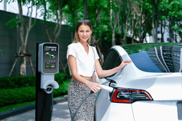 Young woman travel with EV electric car charging in green sustainable city outdoor garden in...