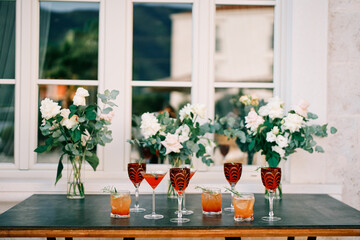 Glasses with alcoholic drinks stand on the table near bouquets of flowers in vases near the window of the building