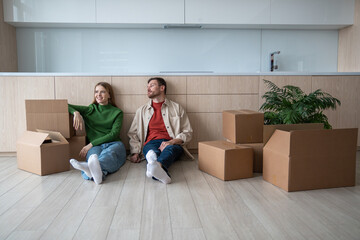 Happy new homeowners tenants renters tired young couple man woman sitting on floor among cardboard...