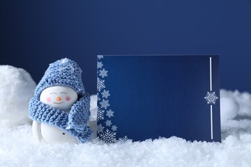 Cute decorative snowman and blank Christmas card on snow against blue background