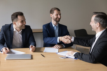 Three satisfied business partners shake hand establish partnership, finish meeting in office conference room. Client and sales managers make deal, handshake, feel satisfied by successful negotiations