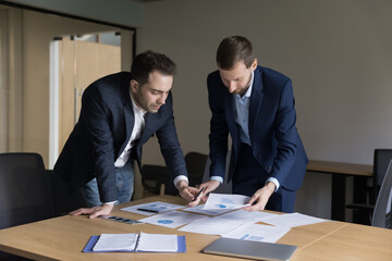 Two business men stand at meeting desk over papers, prepare report, analyze sales, brainstorm on statistic data, talking, work with charts and diagrams, focused employees discuss financial documents