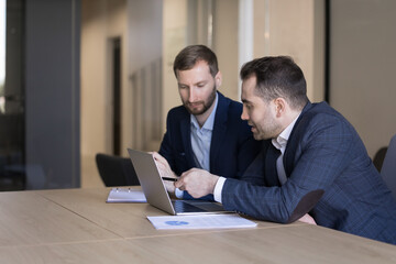 Two young 35s male office employees review joint task, discuss on-line business project use laptop engaged in teamwork, partnership. Manager makes commercial offer to client, met in company boardroom