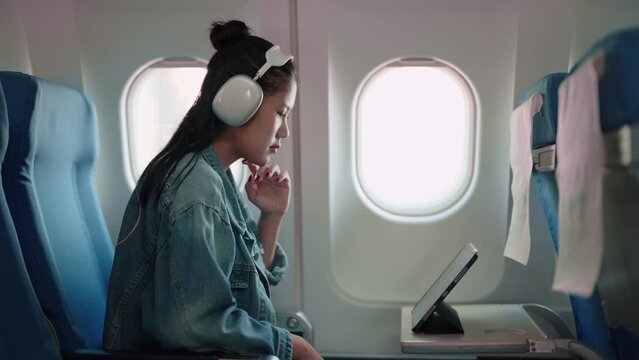 Asian female traveler enjoying a show on her tablet with headphones during the plane journey, anticipating holiday adventures.