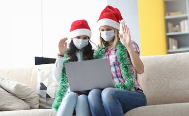 Two woman in protective mask and santa claus hat sit side by side and wave hello. Girlfriend talk...