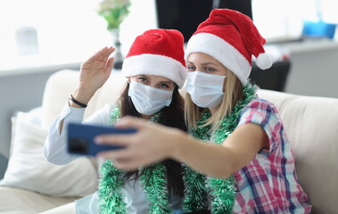 Two women in protective mask and santa claus hat talk online on phone. Girlfriend hold phone in her...